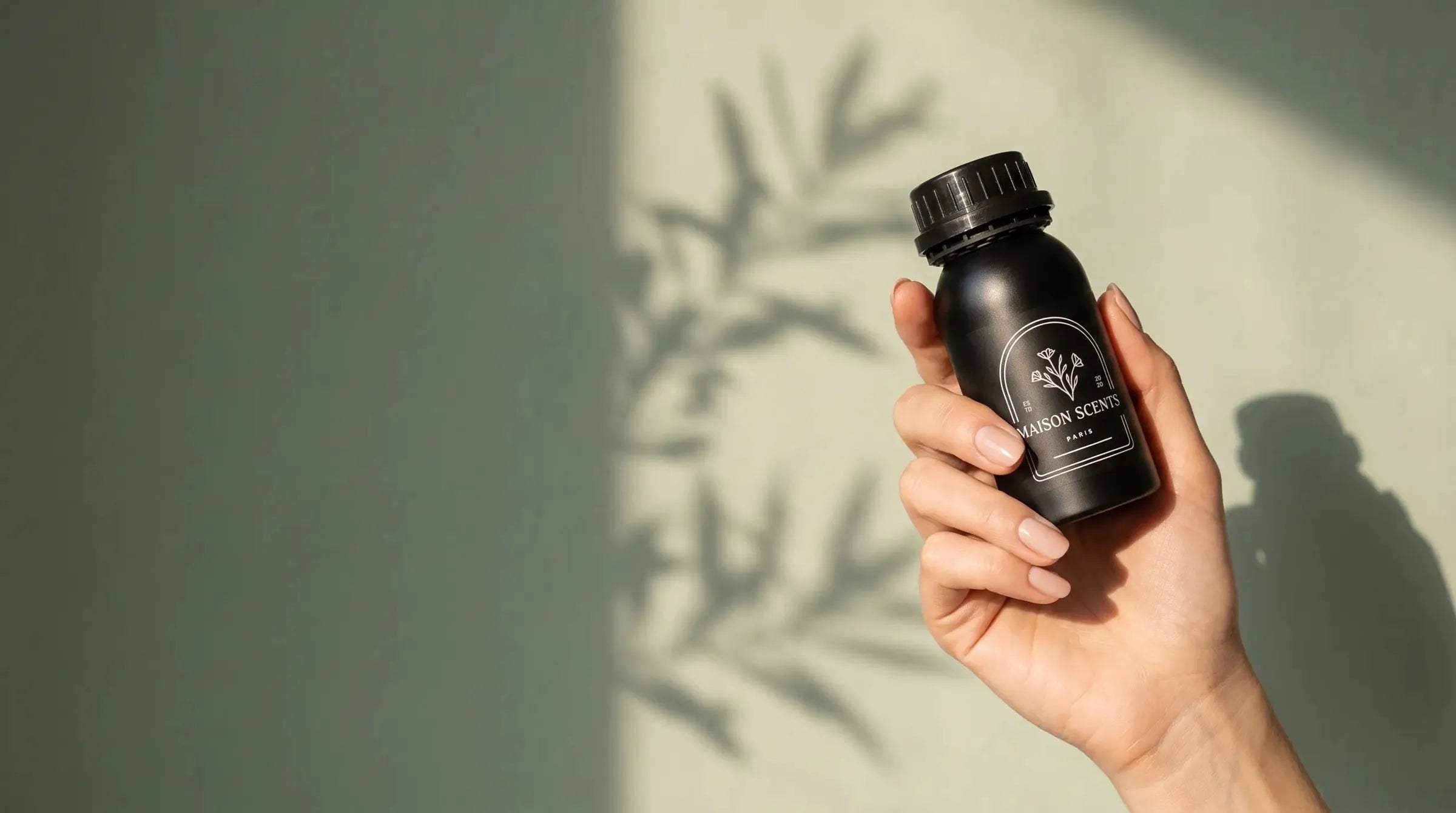 A hand holding The Grand fragrance oil against a sage wall in soft sunlight